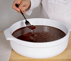 a person is spooning chocolate into a large white bowl on a wooden table with a knife and fork in it