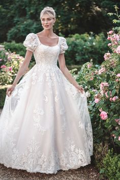 a woman in a white wedding dress standing next to pink flowers and bushes with her hands on her hips