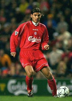 a man in red uniform kicking a soccer ball on field with crowd watching behind him