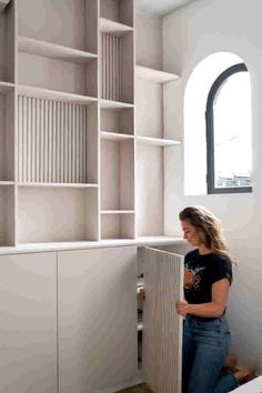 a woman standing in front of a white bookcase next to an open window with shutters