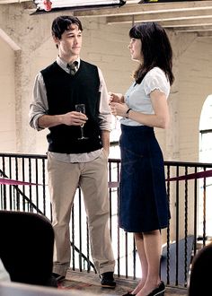 a man and woman standing next to each other in front of a railing holding wine glasses