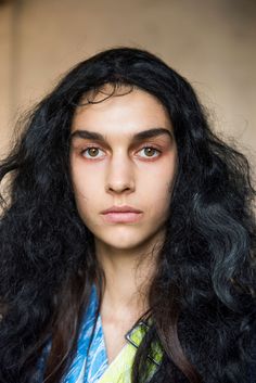 a woman with long black hair and blue shirt looking at the camera while wearing makeup