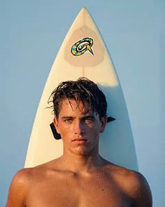 a shirtless young man standing next to a surfboard on the beach with his head tilted back