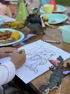 a child is drawing on paper at a table with other toys and plates in the background