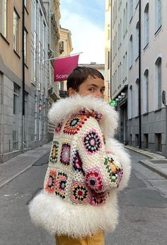 a woman is walking down the street wearing a white jacket with multicolored crochet