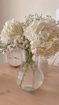 white flowers in a clear vase on a wooden table next to a clock and lamp