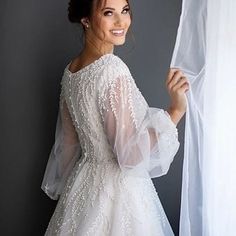 a woman in a white dress standing next to a window with her hands on the curtain