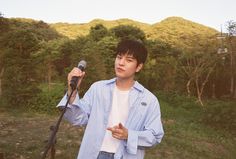 a young man holding a microphone in front of a mountain with trees and bushes behind him