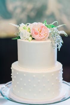 a white wedding cake with pink flowers and greenery on top is sitting on a glass plate