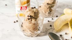 two glasses filled with ice cream next to bananas and chocolate chips on a marble counter