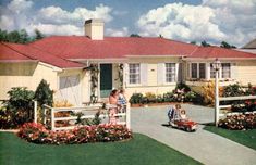 two women and a child are riding a toy car in front of a house with flowers on the lawn