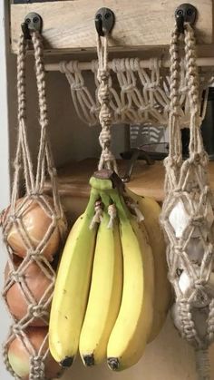 several bananas hanging from hooks on a roped wall with other fruit in the background