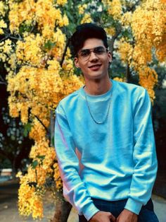 a young man wearing glasses sitting on a bench in front of a tree with yellow flowers