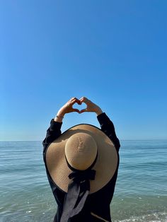 a woman standing on top of a beach next to the ocean holding her hands in the shape of a heart