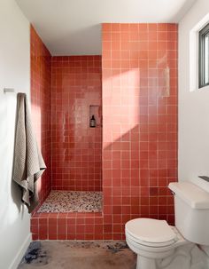 a white toilet sitting next to a walk in shower under a bathroom window with red tiles on the walls