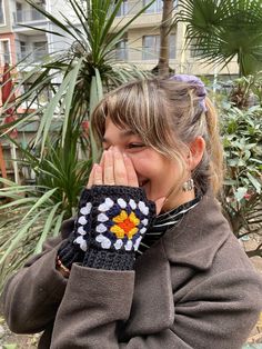 a woman covers her mouth with a hand knitted flowered mitt in front of palm trees