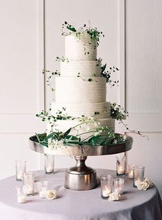 a white wedding cake with greenery on top sits on a silver platter surrounded by candles