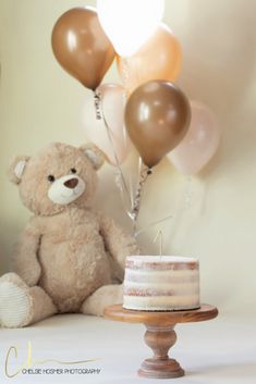 a teddy bear sitting next to a cake with balloons in the air and a candle on top