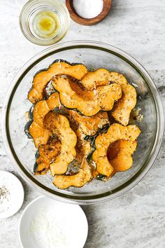 a glass bowl filled with cooked bananas next to bowls of salt and seasoning on a table