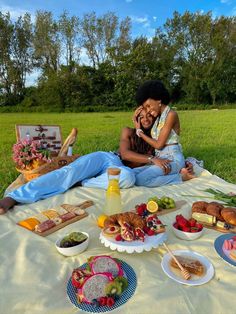 two people sitting on a blanket in the grass with plates of food and drinks around them