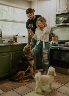 a man and woman are standing in the kitchen with their two dogs looking at something