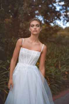 a woman in a white dress is posing for the camera with trees and bushes behind her