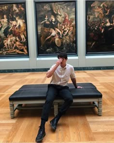 a man sitting on top of a bench in front of some art work with one hand up to his face