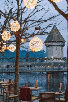 an outdoor dining area with tables and chairs next to the water at dusk, lit up by christmas lights