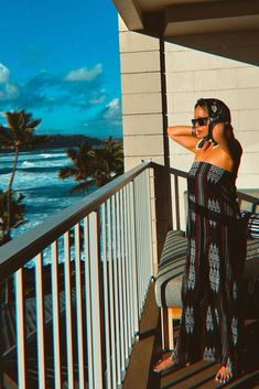 a woman standing on a balcony next to the ocean with her arms behind her head