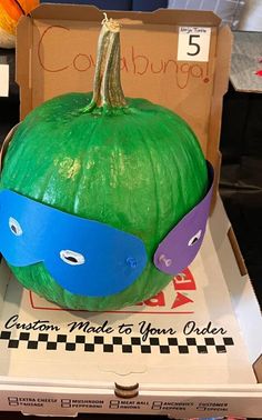 a green pumpkin with a mask on it's head sitting in a cardboard box