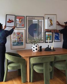 two people standing in front of a table with pictures on the wall behind them and one person pointing at it