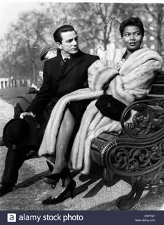 two people sitting on a park bench with fur coats over their shoulders, one man is wearing a suit and the other has a tie