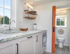 a kitchen with white walls and wood flooring next to a window that has an orange door