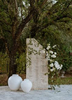 two white vases sitting on top of a table next to a tree and flowers