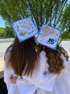 two girls in white robes are facing each other with graduation caps on their heads and trees behind them