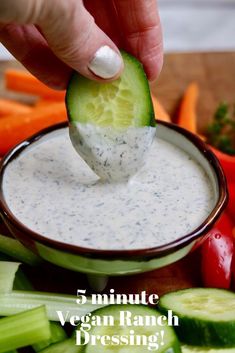 a hand dipping a piece of cucumber into a bowl of ranch dressing surrounded by vegetables