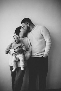 a man and woman kissing while holding a baby in front of a white wall with their hands on each other's heads