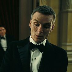 a man in a tuxedo looks at his watch while another man stands behind him