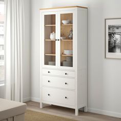 a white cabinet with glass doors and drawers in a room next to a window on the wall