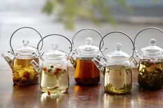 five glass teapots filled with different types of tea sitting on a wooden table