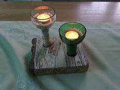 two lit candles are sitting on a piece of wood with a green glass cup next to it