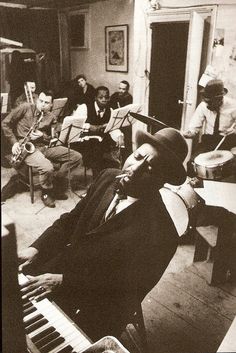 black and white photograph of people playing musical instruments in a room with piano, piano keyboard, and man sitting on chair