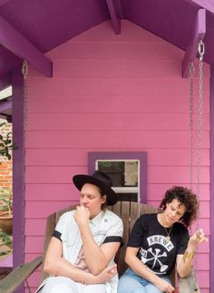 two people sitting on a porch swing in front of a pink house with purple trim