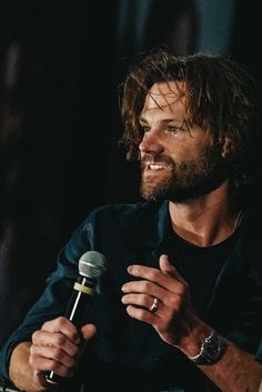 a bearded man holding a microphone in his right hand and looking at the camera while sitting down