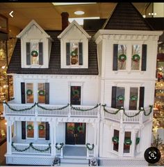 a large white doll house with wreaths and lights on the front windows is displayed in a store window