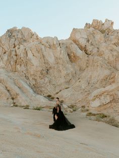 a woman in a black dress sitting on top of a rock covered field next to a mountain