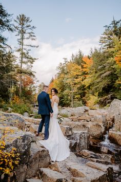 a bride and groom standing on rocks in front of trees with fall foliage around them