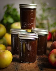 four jars filled with pickles sitting on top of a wooden table next to apples