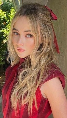 a woman with long blonde hair wearing a red dress and posing for a photo in front of a tree