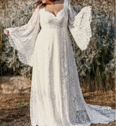 a woman in a long white dress standing on a dirt road with her arms outstretched